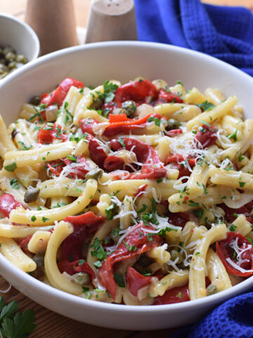 A white bowl with pasta, red peppers and capers.